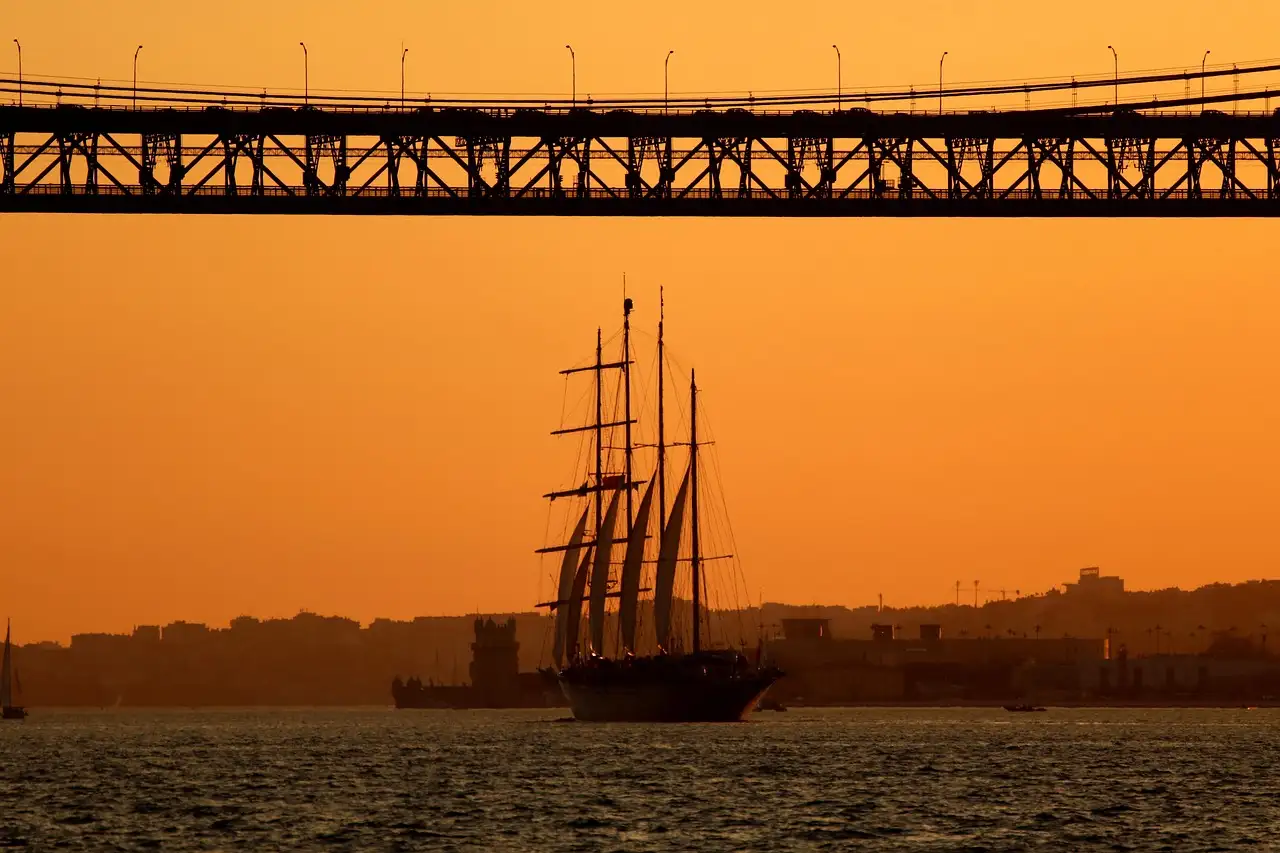 Tall ship silhouetted on the Tagus under the 25 de Abril Bridge in an orange Lisbon sunset