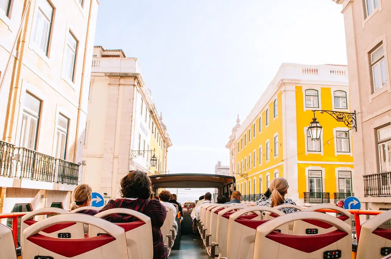 Rows of red seats on the open top deck of a sightseeing bus, with passengers seen from behind, driving past pale and yellow buildings with iron balconies in Lisbon