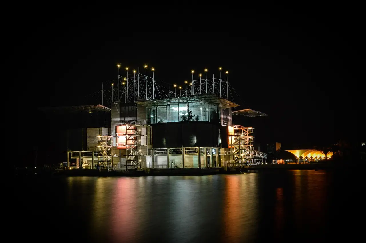 The glass and steel Oceanário de Lisboa at night, lit from within and reflected in the dark water of the dock in Parque das Nações