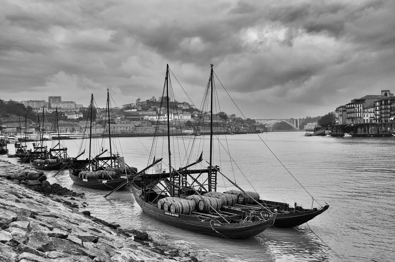 Black and white view across the Douro river in Porto with moored rabelo boats carrying wine barrels, hillside houses opposite and a bridge under a heavy sky