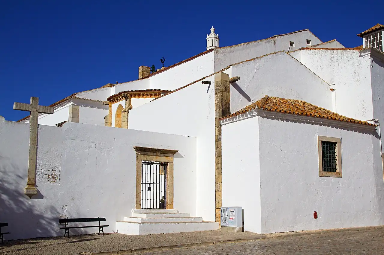 A whitewashed church with a stone cross and an orange-tiled roof under a deep blue sky in the old town of Faro