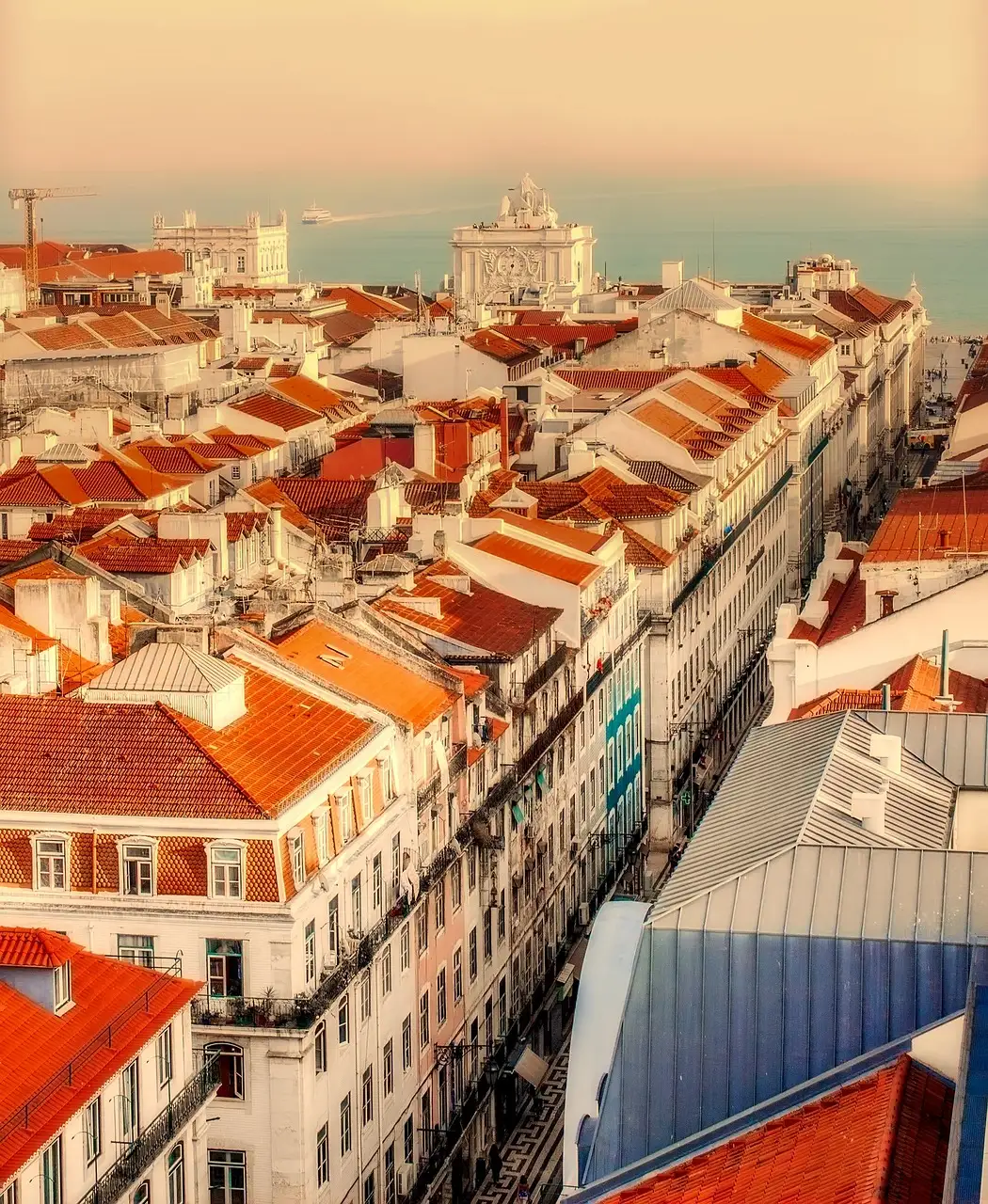 Orange-tiled roofs and tall white buildings in central Lisbon seen from above, with the Rua Augusta Arch at the end of a straight street and the river beyond