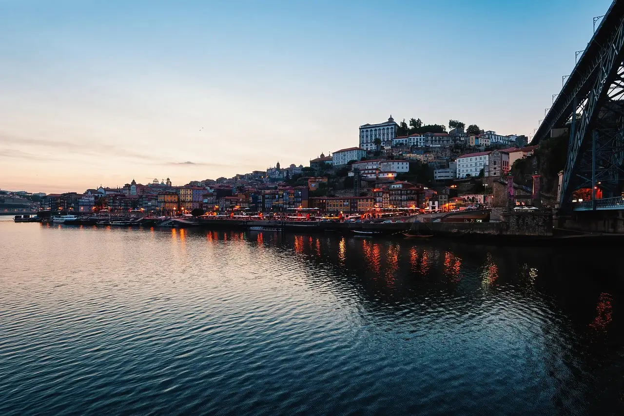 The Douro river in Porto at dusk, with the lit Ribeira waterfront and old town on the hillside beside the iron Dom Luís I bridge