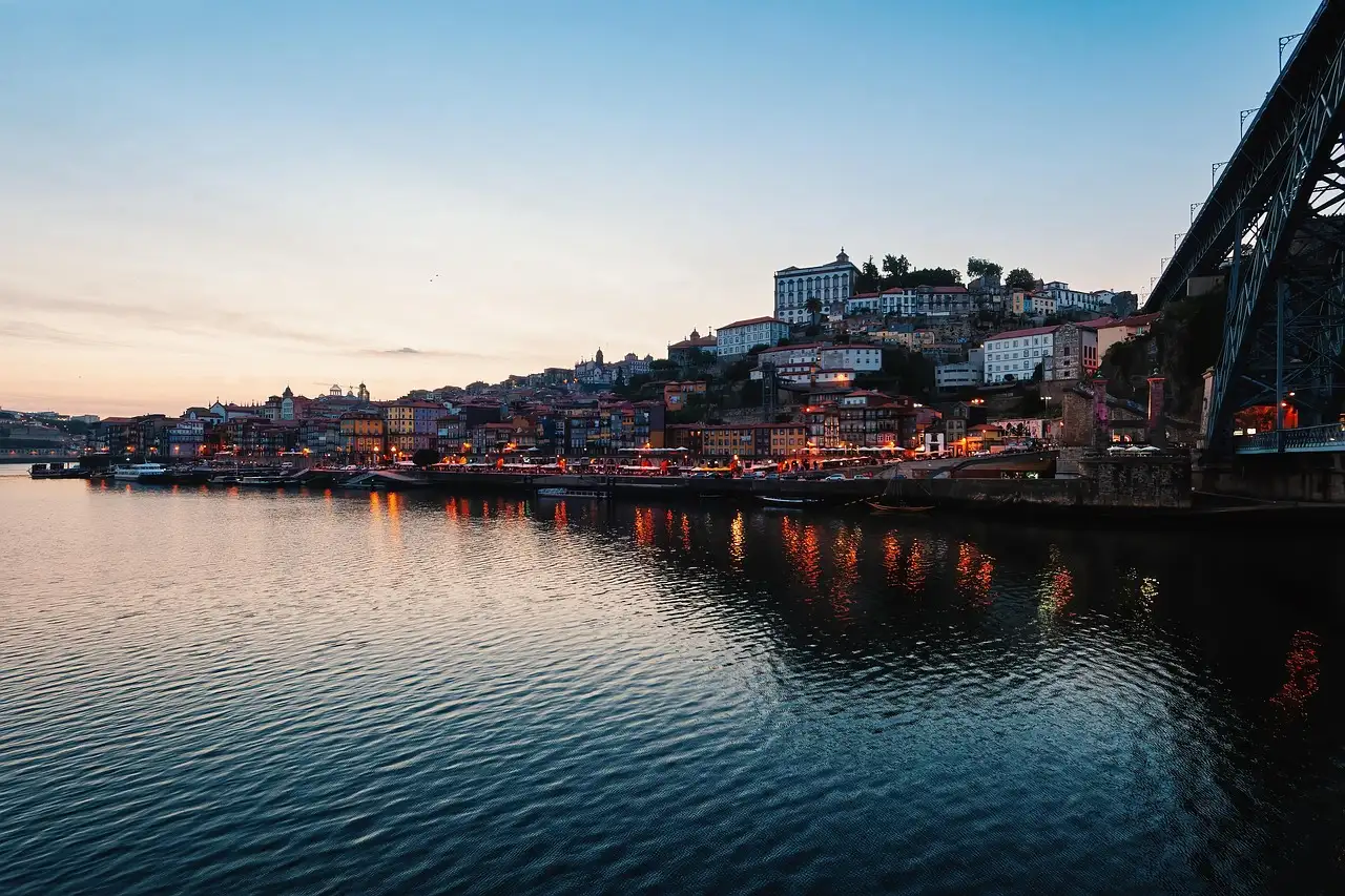 The Douro river in Porto at dusk, with the lit Ribeira waterfront and old town on the hillside beside the iron Dom Luís I bridge