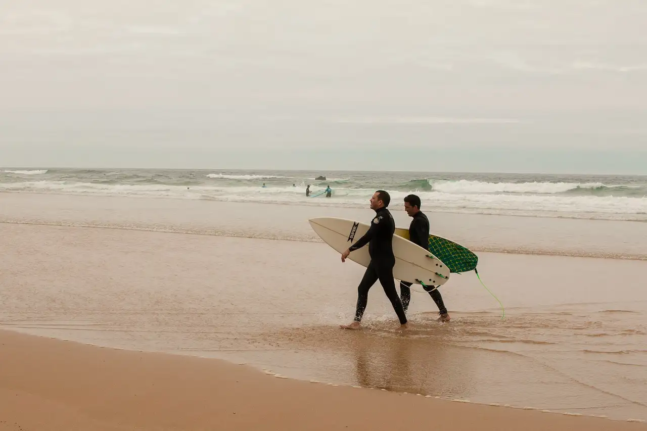 Two surfers in black wetsuits carrying surfboards across wet sand towards the waves on an overcast Portuguese beach