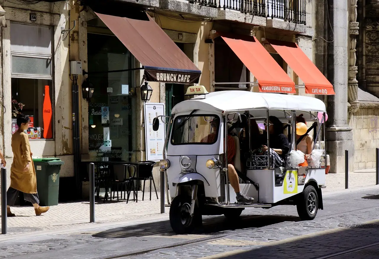 A white three-wheeled tuk-tuk with a canopy and passengers on board, stopped on a cobbled street in Lisbon's old town beside café awnings