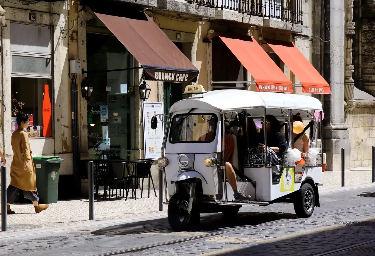 A white three-wheeled tuk-tuk with a canopy and passengers on board, stopped on a cobbled street in Lisbon's old town beside café awnings