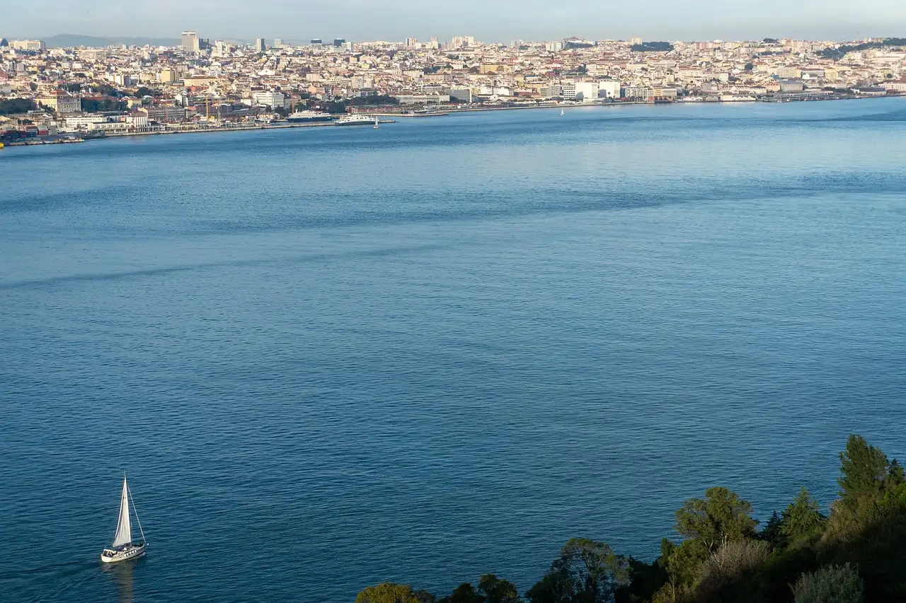 A small white sailboat on the wide blue Tagus with the Lisbon skyline across the water