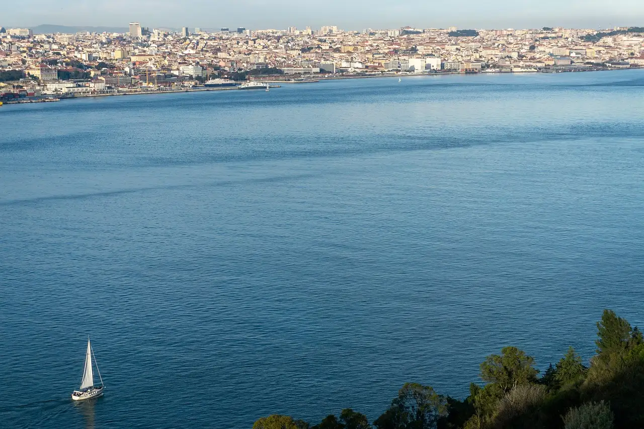 A small white sailboat on the wide blue Tagus with the Lisbon skyline across the water