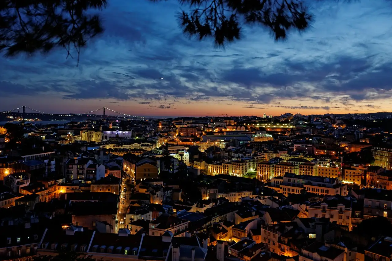 Lisbon's rooftops and streets lit at dusk under a dark blue sky with orange glow on the horizon, the 25 de Abril Bridge on the left