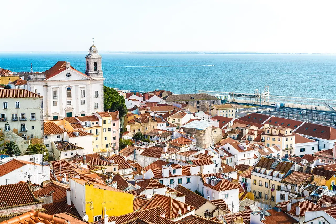 Orange-tiled roofs and pastel houses of Alfama in Lisbon, with a white church and bell tower on the left and the wide blue Tagus river behind
