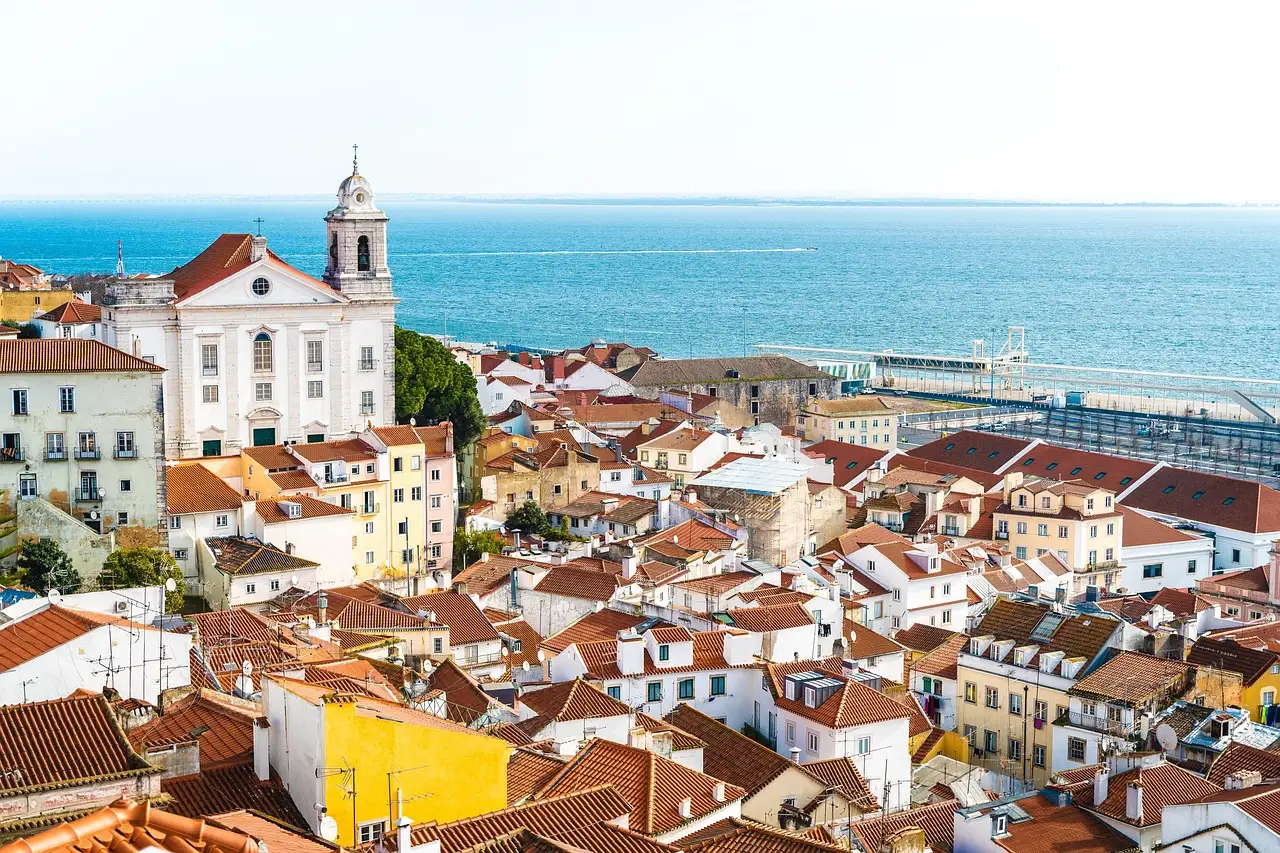 Orange-tiled roofs and pastel houses of Alfama in Lisbon, with a white church and bell tower on the left and the wide blue Tagus river behind