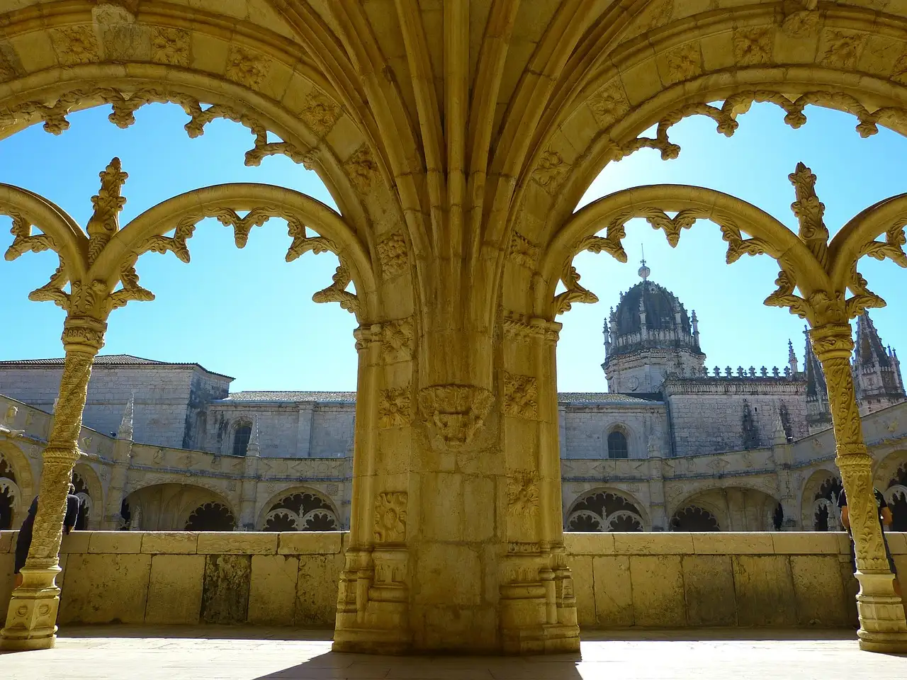 Pointed stone arches and carved columns of the cloister of Jerónimos Monastery in Belém, with the monastery's dome and roofs seen through the arches under a blue sky