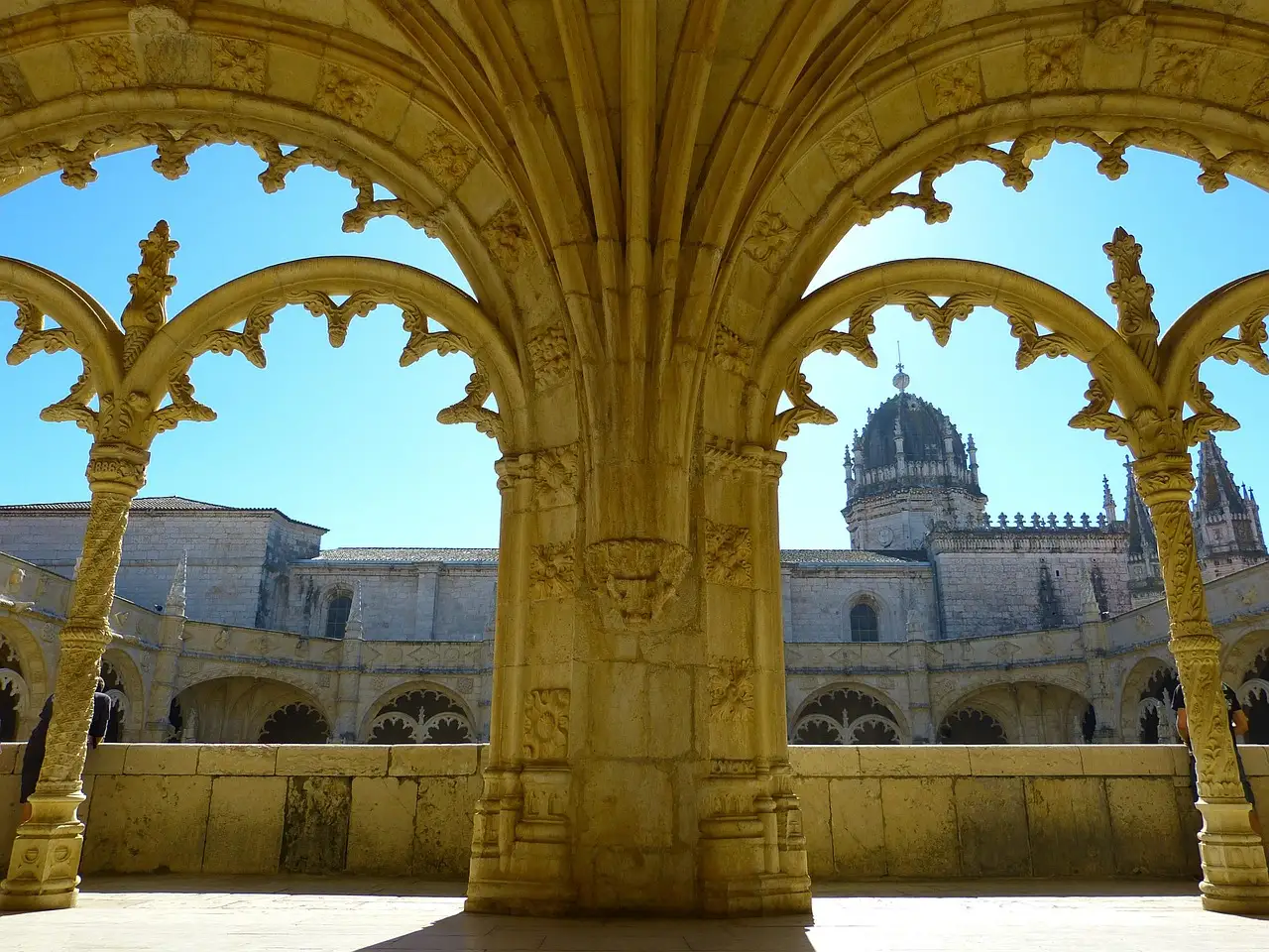 Pointed stone arches and carved columns of the cloister of Jerónimos Monastery in Belém, with the monastery's dome and roofs seen through the arches under a blue sky