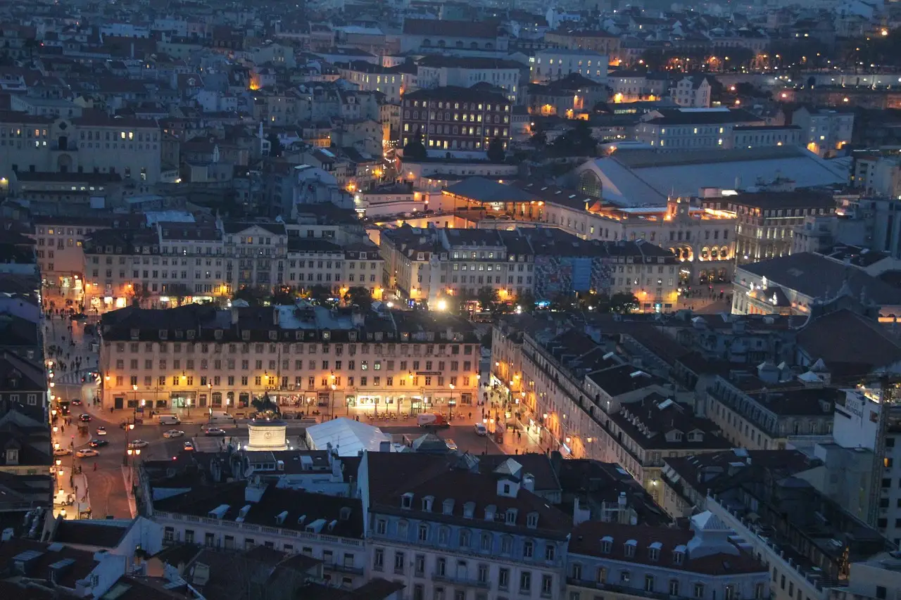 Lisbon's lower town from above at dusk, with street lamps and lit windows coming on along the main squares and streets between the rooftops