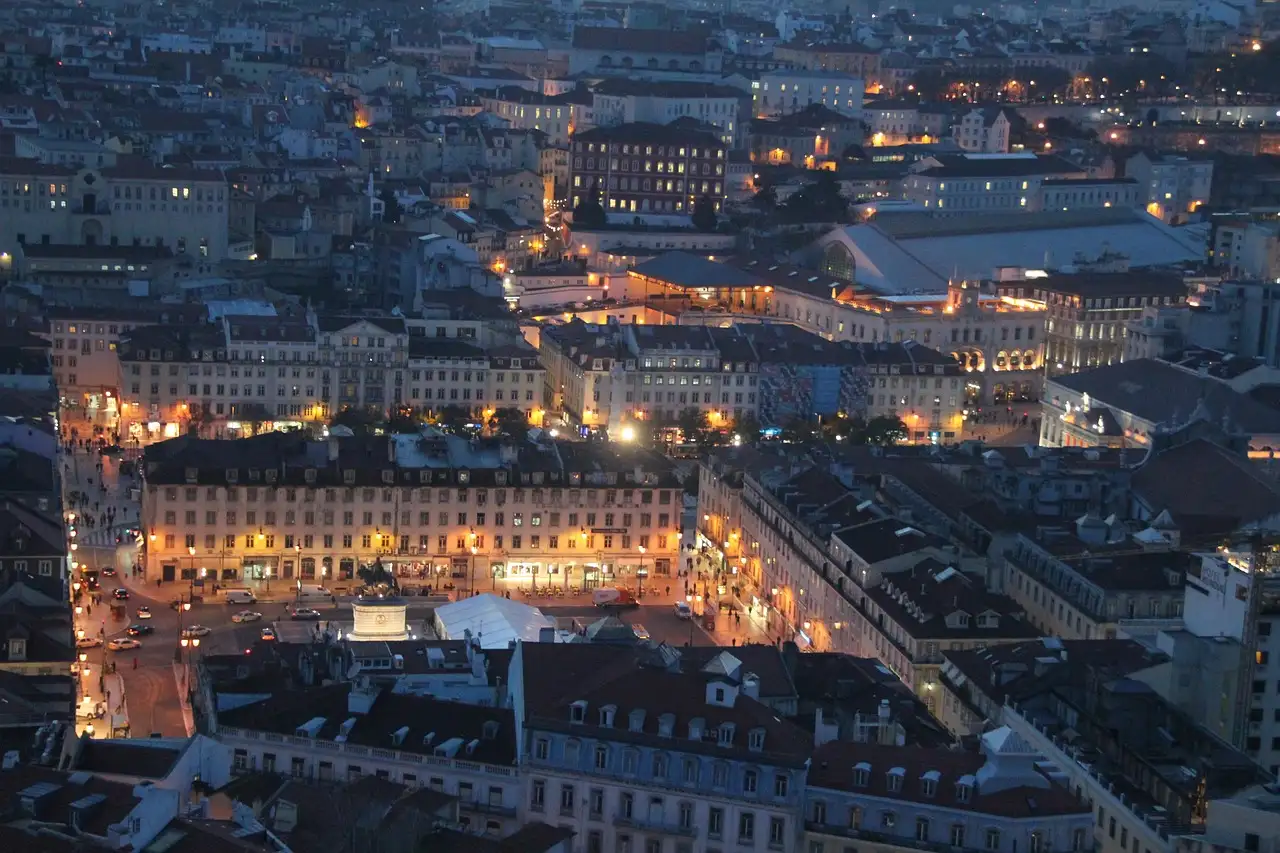 Lisbon's lower town from above at dusk, with street lamps and lit windows coming on along the main squares and streets between the rooftops
