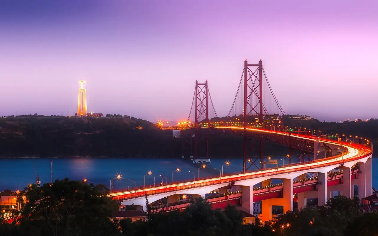 A long road bridge curving over the Tagus river at dusk, with its red towers lit and a tall illuminated statue on the far hill under a purple sky
