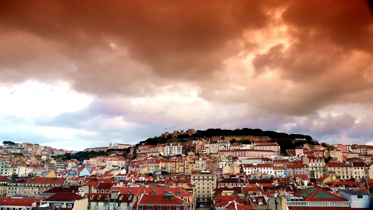The hill of São Jorge Castle rising above the pastel houses of Lisbon's old town under a heavy orange and grey sky