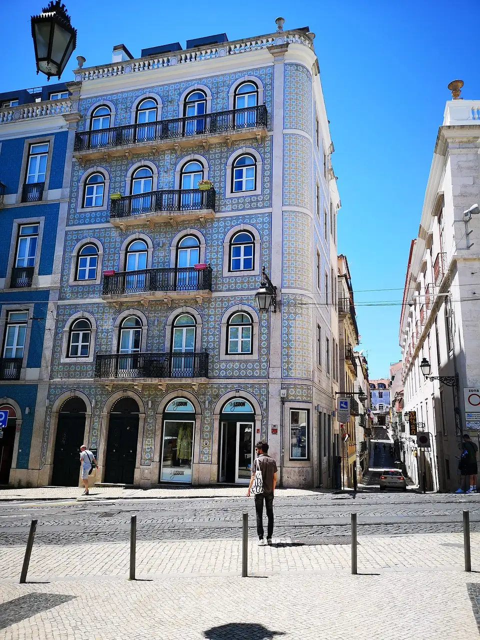 A lone pedestrian crossing a cobbled street in front of a blue-tiled azulejo corner building with arched windows and iron balconies in Lisbon