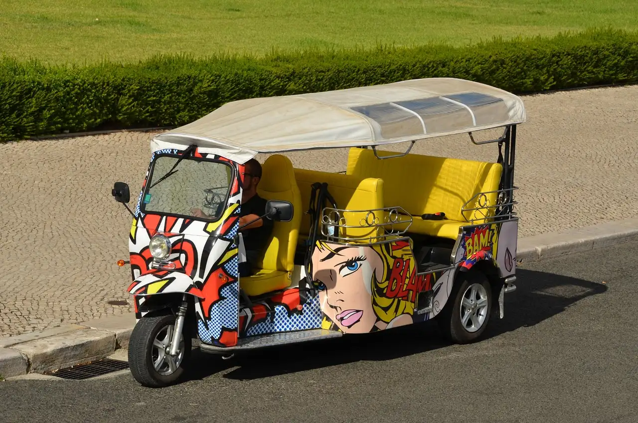 Pop-art painted yellow tuk-tuk parked at the roadside in Lisbon beside a green hedge