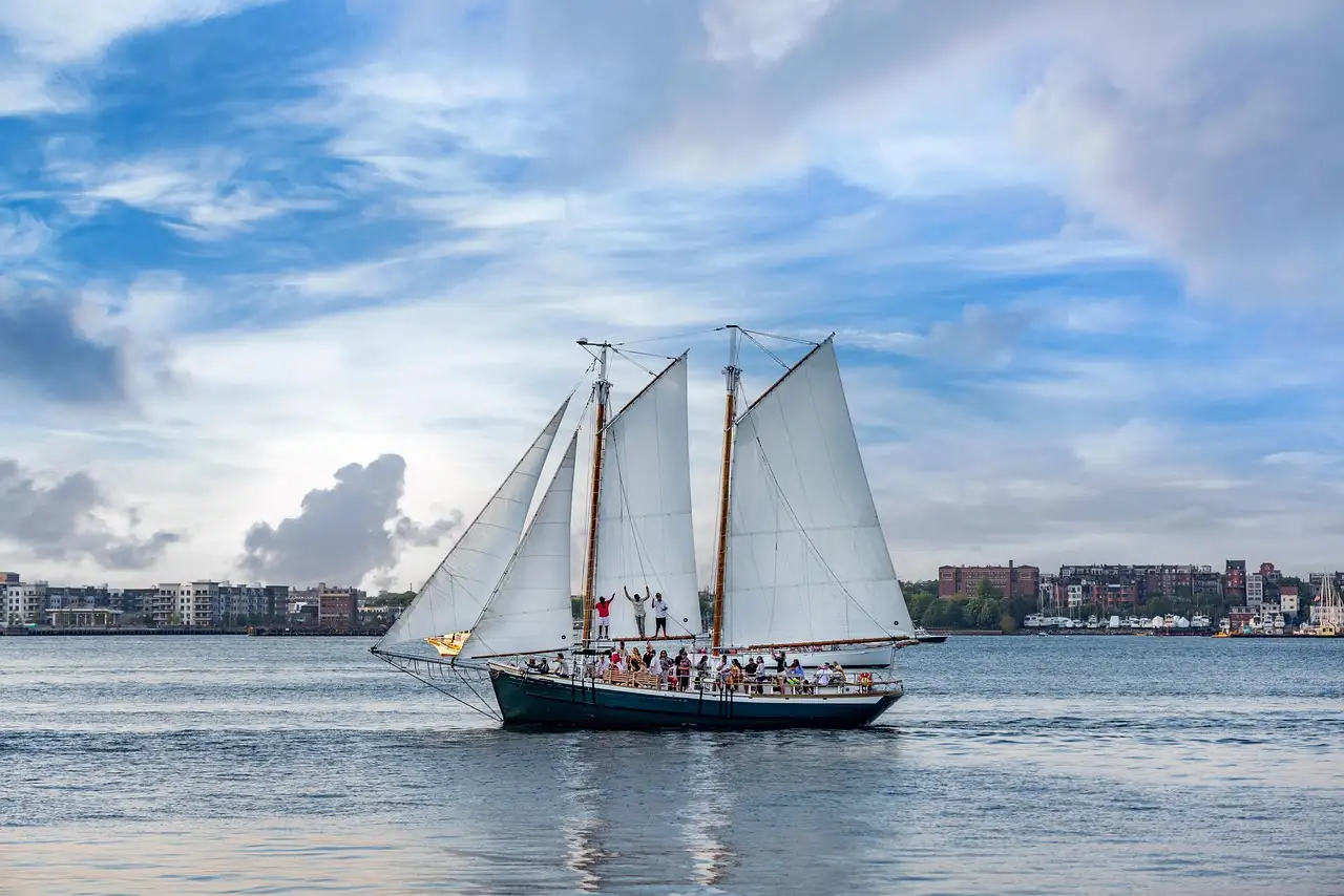 Schooner with white sails crossing a broad river, with a low waterfront behind