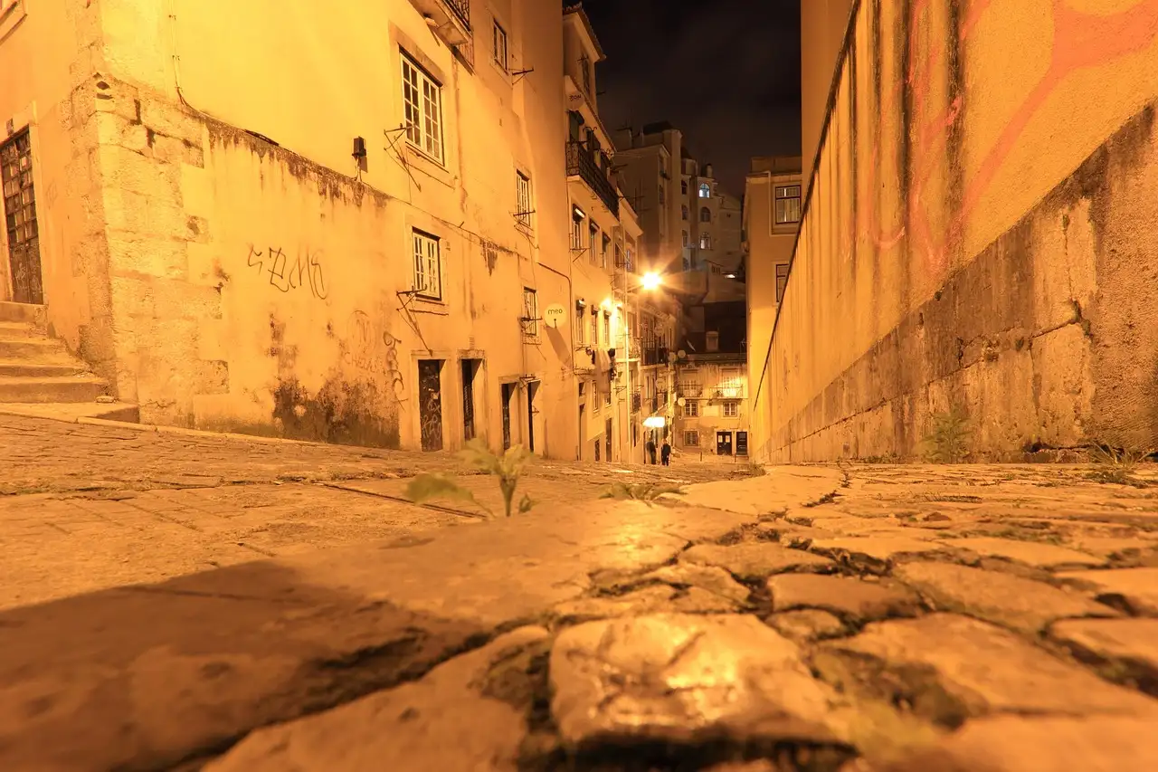 Narrow old-town lane in Lisbon lit warm orange at night, with cobbles and stone walls