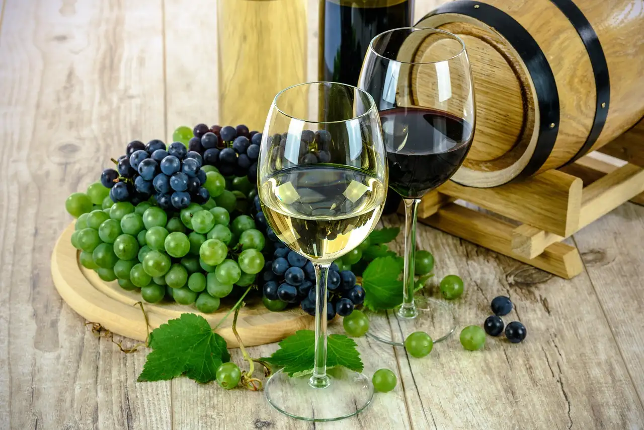 Glasses of white and red wine with grapes and a small barrel on a rustic wooden table