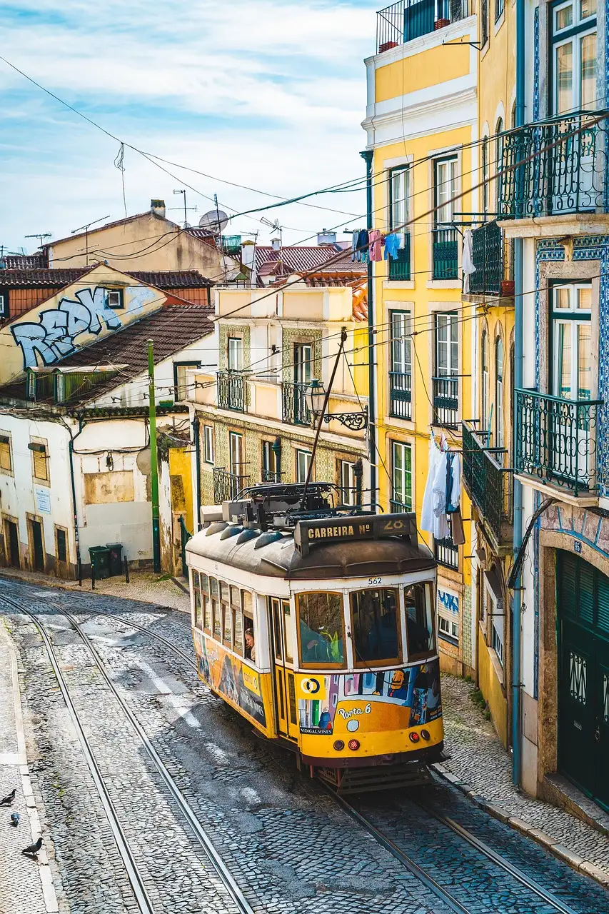 A small yellow tram with a route 28 sign climbing a cobbled street between yellow and white houses with iron balconies in Lisbon